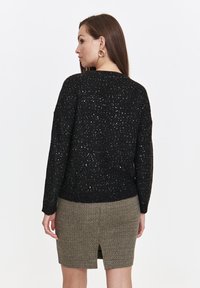 Black knit sweater with a speckled texture, featuring a loose fit and ribbed hem, paired with a check-patterned skirt and minimalistic earrings.