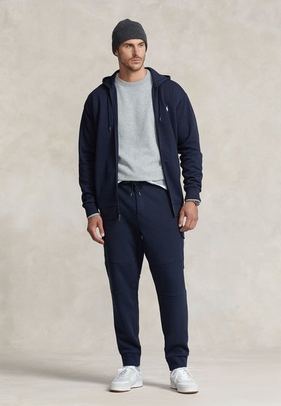 Námořnická mikina s kapucí přes hlavu s šedým svetrem pod ní, doplněná námořnickými tepláky se stahovací šňůrkou a bílými teniskami.