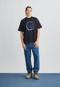 Svart bomulls-T-skjorte med et sirkulært grafisk design i lilla. Brukes med blå denim-bukser og lysegrå ankelstøvler med teksturert finish.