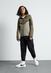 Olivgrüner Reißverschluss-Hoodie mit schwarzen Akzenten, kombiniert mit schwarzen Jogginghosen und weißen High-Top-Sneakern. Verfügt über eine Reißverschlusstasche am Ärmel.