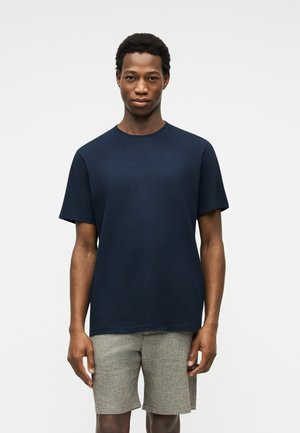 Hombre de pie contra un fondo liso, vestido con camiseta azul marino de mangas cortas y pantalones cortos gris claro hasta la rodilla.