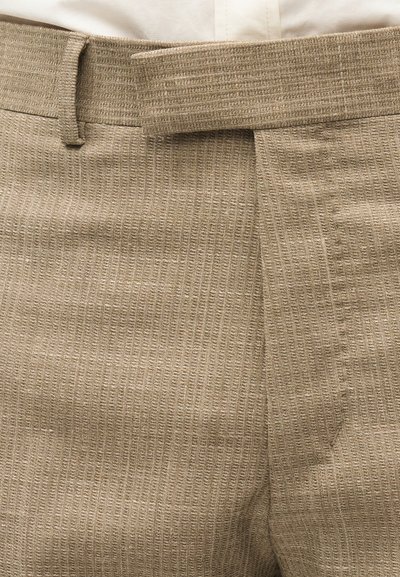 Μπεζ παντελόνι με υφή υφάσματος, συνοδευόμενο από ταιριαστή θηλιά για ζώνη και ορατό κρεμ πουκάμισο που είναι μπατισμένο στη μέση.