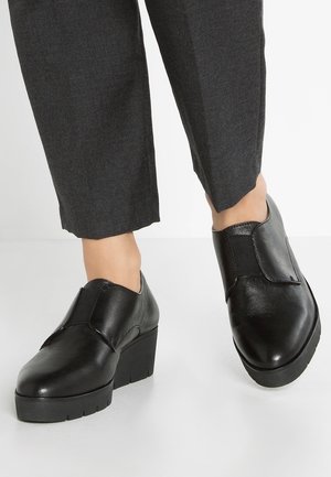 Czarne skórzane buty na nieco podwyższonym obcasie, charakteryzujące się eleganckim designem, elastycznymi bocznymi panelami i matowym wykończeniem. W zestawie z szarymi spodniami.