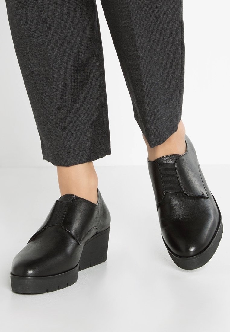 Chaussures en cuir noir avec un talon légèrement surélevé, présentant un design épuré, des panneaux latéraux élastiques et une finition mate. Associées à un pantalon gris.