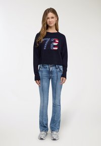 Maglione blu navy lavorato a maglia con design corto, caratterizzato da una grande grafica multicolore "72". Abbinato a jeans a zampa d'elefante blu chiaro e sneakers argento.