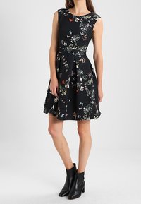 Robe noire sans manches avec imprimé floral multicolore, jupe mi-longue, portée avec des bottines noires à talons par une femme debout.