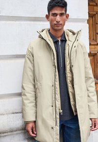 Lys beige pufferjakke med hætte, lynlås og trykknapper. Har sidelommer og en afslappet pasform over en mørkegrå top og denimjeans.