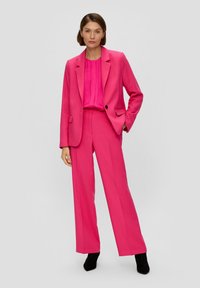 Fuchsie Maßanzugjacke und weit geschnittene Hose aus strukturiertem Stoff, mit einer plissierten Bluse darunter. Schwarze Knöchelstiefel runden das Outfit ab.