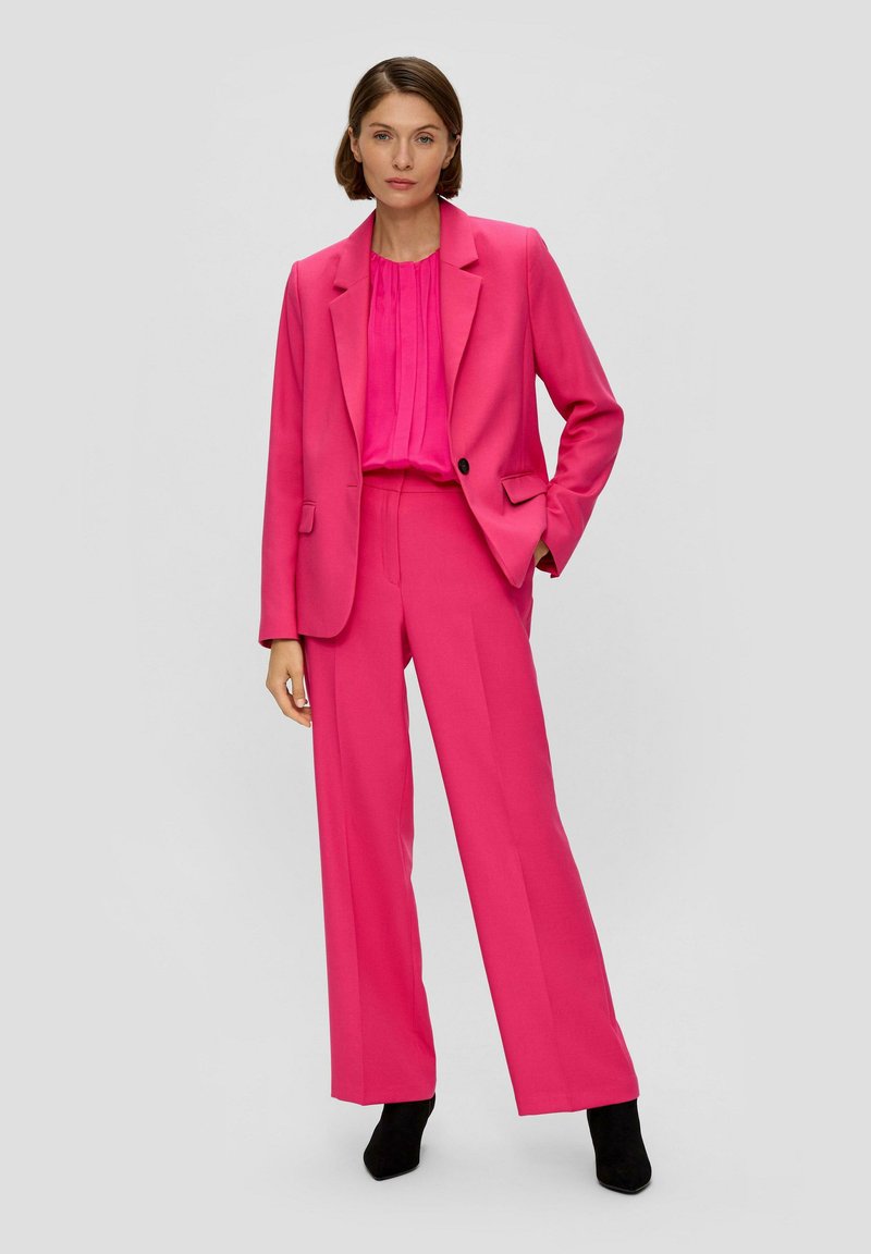 Fuchsie Maßanzugjacke und weit geschnittene Hose aus strukturiertem Stoff, mit einer plissierten Bluse darunter. Schwarze Knöchelstiefel runden das Outfit ab.