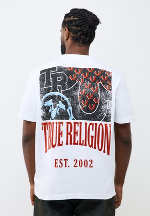 Mann mit geflochtenem Haar, der ein weißes T-Shirt mit großem buntem True Religion-Logo und Text "EST. 2002" auf dem Rücken trägt.