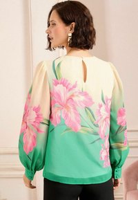 Blusa floreale con fiori rosa e verdi su uno sfondo crema. Maniche lunghe e voluminose con polsini abbottonati e un dettaglio a chiave sulla schiena.