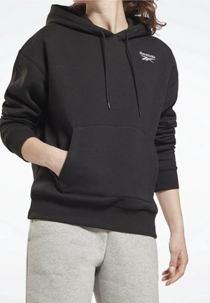 Felpa nera con cappuccio, tasca frontale, cappuccio con cordini regolabili e logo Reebok. Vestibilità casual con polsini e orlo a coste. Indossata con pantaloni da tuta grigi.