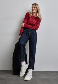 Rot gemusterter Strickpullover, dunkelblaue High-Waist-Jeans, weiße Sneakers mit grauen Akzenten und eine schwarze Jacke über einem Arm. 