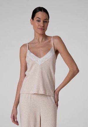 Donna con top beige senza maniche con bordatura in pizzo bianco e pantaloni coordinati, in piedi davanti a uno sfondo grigio uniforme.