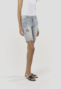 Világos kék farmer bermuda rövidnadrág virágos hímzéssel és díszítő folttal, felhajtott szegéllyel és elülső zsebekkel.