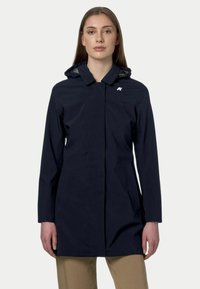 Manteau imperméable marine avec capuche, manches longues, fermeture éclair frontale et poches latérales. Présente un petit logo sur la poitrine. Texture lisse.