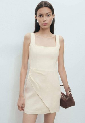 Robe ajustée crème avec décolleté carré, bretelles larges et détail drapé. Mannequin tenant un petit sac à main marron. Texture de tissu lisse.