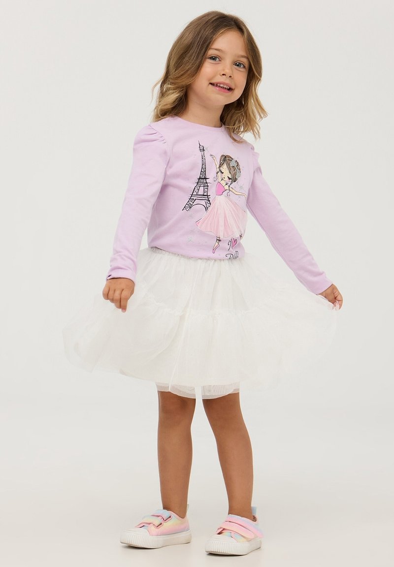 Maglietta a maniche lunghe di un viola chiaro con un design illustrato, abbinata a una gonna di tulle bianca a strati e sneakers dai colori pastello.