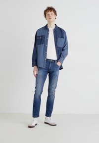 Camicia in denim con un design a blocchi di colore blu chiaro e scuro, t-shirt bianca a collo rotondo, jeans blu scuro e sneakers bianche con suole marroni.