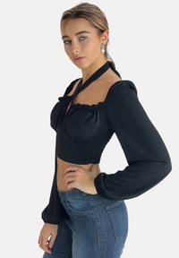 Zwarte cropped blouse met opgeblazen mouwen, verstelbare strikjes bij de halslijn en een gestructureerde stof. Gecombineerd met nauwsluitende blauwe jeans.
