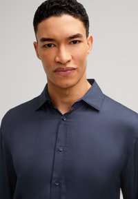 Chemise bleu marine à boutons avec une texture lisse et un col pointu. Comprend six boutons et une coupe ajustée, conçue pour un look soigné.