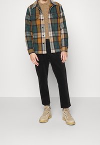 Osoba ve hnědém pleteném svetru, vícebarevné zeleno-hořčicové kostkované bundě, černých kalhotách a béžových šněrovacích botách stojící na světlé podlaze.