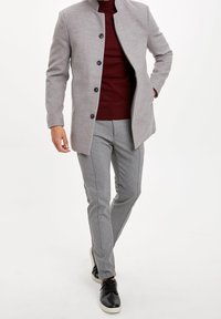 Grauer Wollmischmantel mit Stehkragen und Knopfleiste; darunter ein burgunderroter Strickpullover; graue Slim-Fit-Hose; schwarze Sneakers vervollständigen das Outfit.