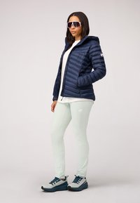 Marineblauwe pufferjack met capuchon en ritszakken, gecombineerd met lichtgroene leggings met naden en een logo. Donkere schoenen met blauwe accenten.