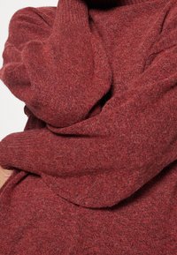 Primo piano di una persona che indossa e abbraccia un maglione rosso lavorato a maglia, con tessuto spesso e testurizzato e maniche lunghe.