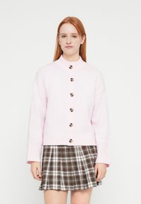 Gaiši rozā adīts kardigans ar augstu apkakli, sešām lielām brūnām pogām priekšpusē un rievotiem manžetēm. Kombinēts ar brūnu rūtainu svārku.