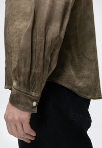 Poignet de chemise en daim marron à manches longues avec bouton sur le bras d'une personne suspendu à côté de la poche d'un pantalon noir.