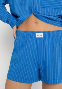 Blå randiga shorts med elastiskt midjeband och en vit etikett med texten "LSCN." Tyget är lätt och mjukt.