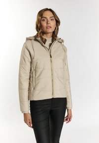 Jachetă puffer bej, matlasată, cu glugă, fermoar aurie și detalii cu șnur, asortată cu pantaloni negri mulati. Material neted, ușor.