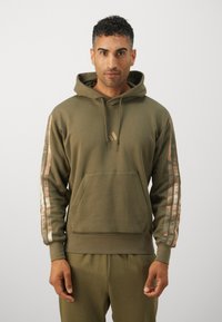 Felpa verde oliva con tasca frontale, cappuccio con cordini e dettagli decorativi lungo le maniche. Il tessuto appare morbido e strutturato.