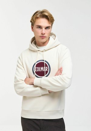 Junger Mann mit verschränkten Armen, der einen cremefarbenen Hoodie mit einem roten und marineblauen runden "COLMAR" Logo auf der Brust trägt.