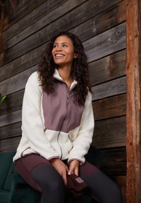 Veste en polaire avec un haut marron clair et un bas crème, fermeture éclair sur le devant, col haut et poches latérales. Modèle assis avec un legging noir et un bonnet en tricot.