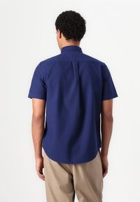 Camicia a maniche corte in cotone blu navy. Presenta un colletto punta, dettagli con piega sul retro e orlo curvo. Indossata con pantaloni beige.