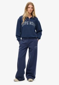 Mørkeblå zip-up sweatshirt med "SUPERDRY" i fede grå bogstaver, parret med matchende løstsiddende sweatpants med "BOSTON" detalje.
