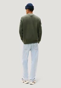 Maglione verde lavorato a maglia con polsini e orlo a coste, abbinato a jeans azzurri. Il modello indossa un berretto lavorato a maglia blu e scarpe da ginnastica casual.