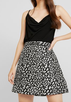 Frau trägt ein schwarzes ärmelloses Oberteil und einen schwarz-weißen Leopardenmuster-Rock, steht mit einer Hand in der Hüfte.