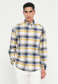Junger Mann, der ein kariertes Hemd in Gelb, Marineblau und Weiß mit Kragen und weiße Hosen trägt, vor einem schlichten weißen Hintergrund steht.