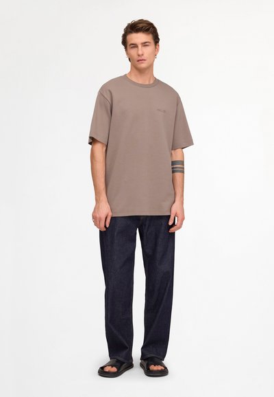 Jeune homme aux cheveux courts portant un t-shirt oversize taupe, un jean sombre ample, des sandales noires et un tatouage à trois bandes sur l'avant-bras gauche.
