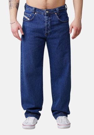 Mann trägt lockere dunkelblaue Jeans und weiße Turnschuhe, steht mit entspannten Händen neben dem Körper vor einem schlichten Hintergrund.