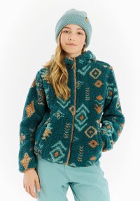 Giacca in pile color teal con dettagli a motivo arancione e blu, dotata di zip frontale e colletto alto. Indossata con un berretto lavorato a maglia blu chiaro.