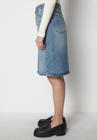 Jupe midi en denim bleu clair avec une coupe droite, poches avant et un ourlet qui tombe juste au-dessus du genou. Associée à des mocassins compensés noirs.