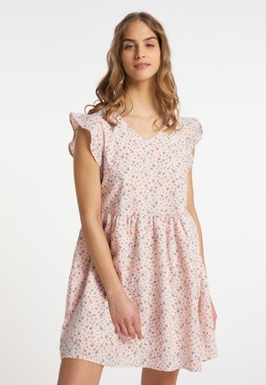 Jonge vrouw met golvend haar, gekleed in een roze bloemenmouwloze jurk met gerimpelde schouders, staand tegen een witte achtergrond.