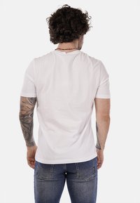 Uomo con capelli ricci, tatuaggi su entrambe le braccia, che indossa una maglietta bianca semplice e jeans blu, in piedi rivolto di spalle su uno sfondo bianco.