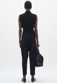 Combinaison noire sans manches avec une coupe ajustée, taille haute et jambes fuselées. Tissu lisse et design minimaliste. Sac noir tenu à la main.