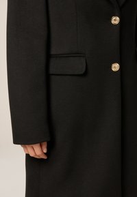 Cappotto nero in tessuto testurizzato, con una tasca sul petto e bottoni dorati. La manica ha un taglio dritto e una finitura pulita.