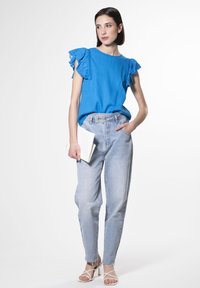 Maglietta blu con maniche a sbuffo, abbinata a jeans a vita alta azzurri e sandali con tacco bianchi. Tiene una pochette bianca.